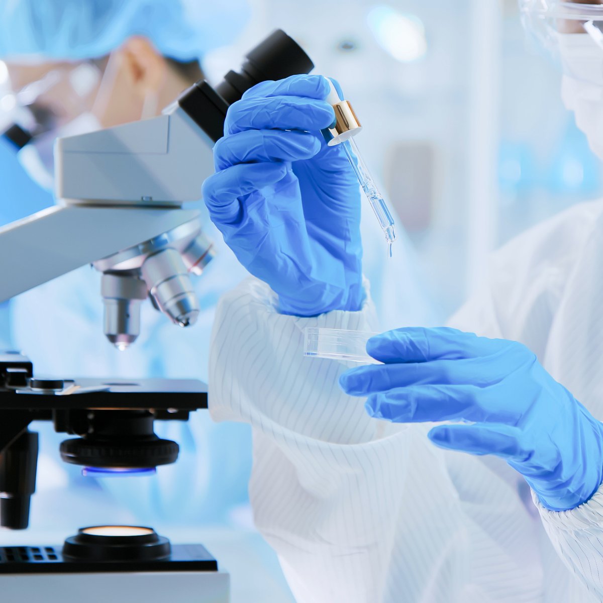 Scientist in a lab coat and gloves examining a dropper of serum beside a microscope