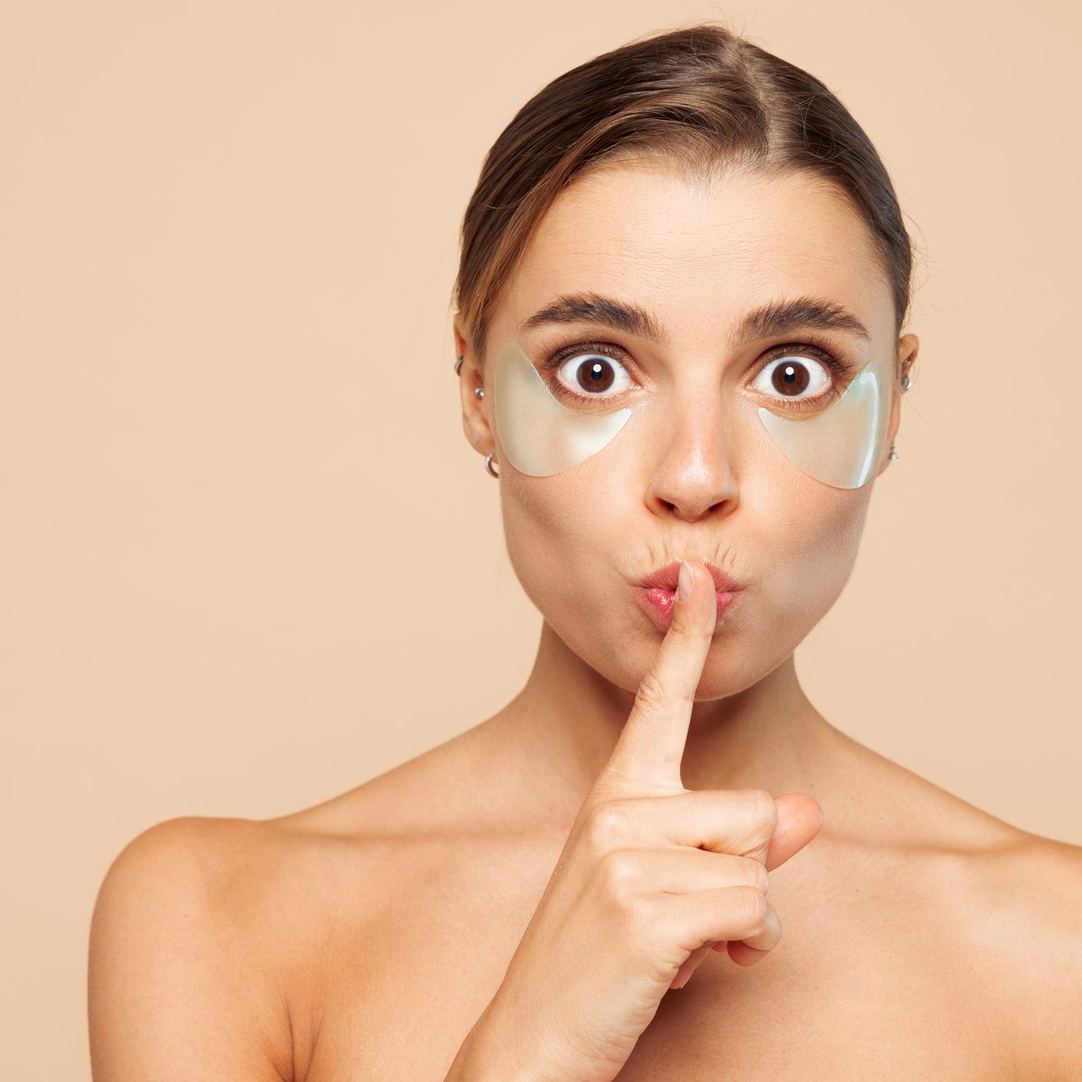 Woman wearing gold under-eye hydrogel patches, finger to her lips, against a soft beige background