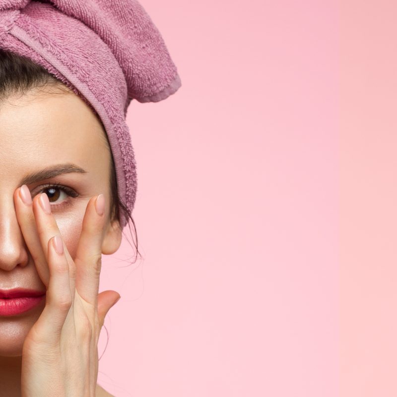 Woman in a pink towel turban resting a hand over one eye against a soft pink background
