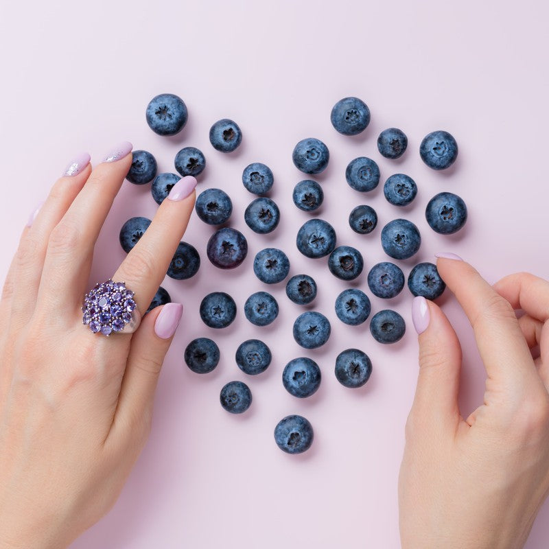 Blueberries arranged in a heart shape on a pink background, framed by manicured hands